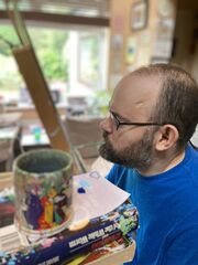 A photo of a man with not much hair and glasses, painting with a paintbrush held in his mouth.