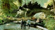 A group of Stegosaurus move into a clearing, watched by a group of humans.