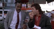 Riggs munches on a hot dog while Murtaugh holds a Pepsi can.