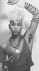 A black and white photo of young RuPaul styled in genderfuck drag. He looks to the camera with a smoldering expression and one arm above his head.