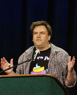 A brown haired man with a light brown plaid shirt stands behind a podium against a black background.