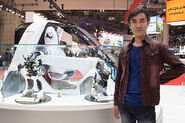 Kawamori standing near a concept car / robot at the Tokyo Motor Show.