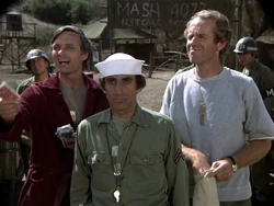 Hawkeye changes some scrip with Winchester. He is handing some new red scrip to Winchester with his right hand. In his left, he has Winchester's old blue scrip which he will then change for new red scrip for himself. He is giving Winchester the same deal - handing over 10 cents of new red scrip for every dollar of old blue. And he is only taking enough to make $400 to repay the money he lost for Boone. B.J. is holding Winchester's bag with the rest of the soon-to-be useless blue scrip.