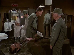 Hawkeye has just called to Carmine by name and asked him to prep a patient. Here he is passing behind. Note the sideburns. This scene is from Season 5 Hepatitis.