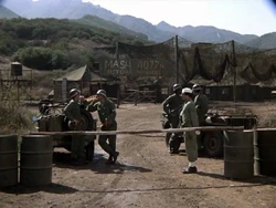 A good view of the camp gate. In the background is the motor pool and the water tower is at the extreme left. To the right, out of the camera view, is the Officers Club. Klinger is on guard, now dressed as a midshipman - he wants to try for the Naval Academy next.