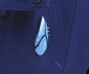 A Starfleet insignia tricom badge, cadet version, with a split arrowhead shape atop a third lower lobe. It lacks the oval surrounding frame seen on the standard tricom badge used by Starfleet officers.