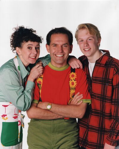 The three cast members stand smiling on a white background publicity shot.