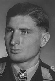 The head of a young man, shown in semi-profile. He wears a military uniform with a military decoration displayed at the front of his shirt collar. His hair is dark and short and combed to his right, his nose is long and straight, and his facial expression is emotionless; looking to the right of the camera.