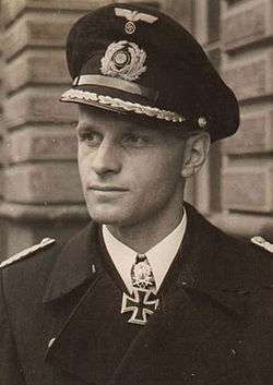 The head and shoulders of a young man, shown in semi-profile. He wears a peaked cap and black naval coat, a white shirt with an Iron Cross displayed at the front of his shirt collar. His facial expression is a determined and confident smile; his eyes gaze into the distance to the left.