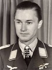 Black-and-white photograph showing the face and shoulders of a young man in uniform. His hair appears dark and is combed to the back. The front of his shirt collar bears an Iron Cross decorations, black with light outline. He is looking at to right of the camera, his facial expression is determined.
