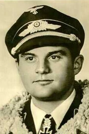 The head and shoulders of a young man, shown in semi-profile. He wears a peaked cap and a pilot's leather jacket with a fur collar, with an Iron Cross displayed at the front of his shirt collar. His nose is long and straight, and his facial expression is a determined and confident smile; his eyes gaze into the distance.