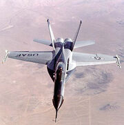 Jet fighter over-flying desert. The jet has black nose and two outward-canted vertical stabilizers between which lay two engines.