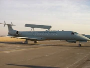 A grey plane with two engines in the rear and a radar on top stays on the ground inside an airfield.