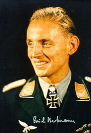 A black and white photograph of a smiling young man wearing a military uniform, peaked cap, various military decorations including a neck order in shape of an Iron Cross.