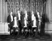 A group pose of eight men in smart evening wear. The King sits in the middle surrounded by his prime ministers.