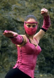 This image shows a masked woman with braided black hair, in a martial arts pose, wearing an a pink-and-black outfit.