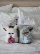A Passionfruit Emoto-Tronic Furby with a Sleepy Blue Emoto-Tronic Furby on a hotel bed.
