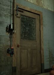 The image shows a plain wooden with a glass square window. To the left side there is a device hooked up to the door, with a battery powered switch at the bottom that is connected via a wire up to a powered wooden plank that is upwards connected to a cross shaped piece of pipe.