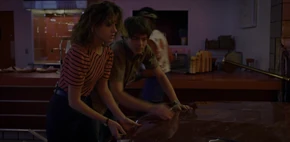 Lucas walking past Nancy and Jonathan as they work to open the car's hood before the Mind Flayer arrives at the mall.