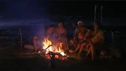 A group of Māori sitting by a fire cook meat.