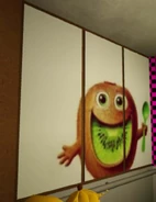 A kiwi with eyes, a slice taken made to resemble a smile, and arms, holding a spoon on the left.