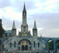 Zicht op de ingang van de Rozenkransbasiliek in Lourdes. Daarboven de torens van de Basiliek van de Onbevlekte Ontvangenis.