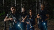 A promotional shot of S.T.A.R.S. members Chris Redfield (Robbie Amell), Richard Aiken (Chad Rook), Jill Valentine (Hannah John-Kamen), and Albert Wesker (Tom Hopper) in the Arklay Forest, likely approaching the crashed helicopter.