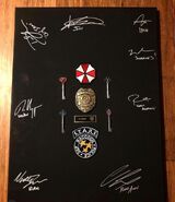 A board containing signatures from Tom Hopper (Wesker), Donal Logue (Irons), Hannah John-Kamen (Jill), Avan Jogia (Leon), Johannes Roberts, Robbie Amell (Chris), Chad Rook (Richard) and Nathan Dales (Brad)