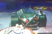 Lizzard has one of its eyes knocked off as it pushes Bamm Bamm into an angle grinder