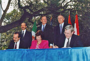 Two men and one women sit at a table and sign a piece of paper, while three men in suits stand behind them, in front of a set of limp flags