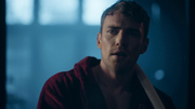 Ben stands alone in a library like room, the background is lit blue to signify it is nighttime. He is dressed in a red robe, and has taken off his bandages. Ben has blonde, short cropped hair, a stubble and blue eyes. He stares back at Jed.