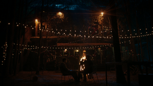 A night time scene in the forest. A large, debilitated wooden house stands central to the scene, with multicoloured fairy lights hanging over it in chains. In the foreground, a fire burns and either side of it are Jed to the left and Ben to the right. They sit comfortably sharing beers.