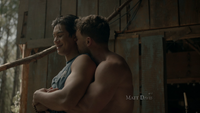 Jed, dressed in a blue sleeveless shirt, is shown waist up standing smiling as Ben, shirtless, embraces him from behind and kisses his neck.