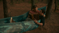 Jed and Ben lie in a blue hammock together, tied between two pine trees in the forest. Jed is dressed in a black long sleeve shirt, rolled up to his elbows, and has a large weathered leather book open in his hand. He is leaning against Ben, who is dressed in a light pink shirt and has his left arm around Jed while his right holds the book too. Jed is looking up at Ben, questioning something in the book, and Ben looks down at him fondly.