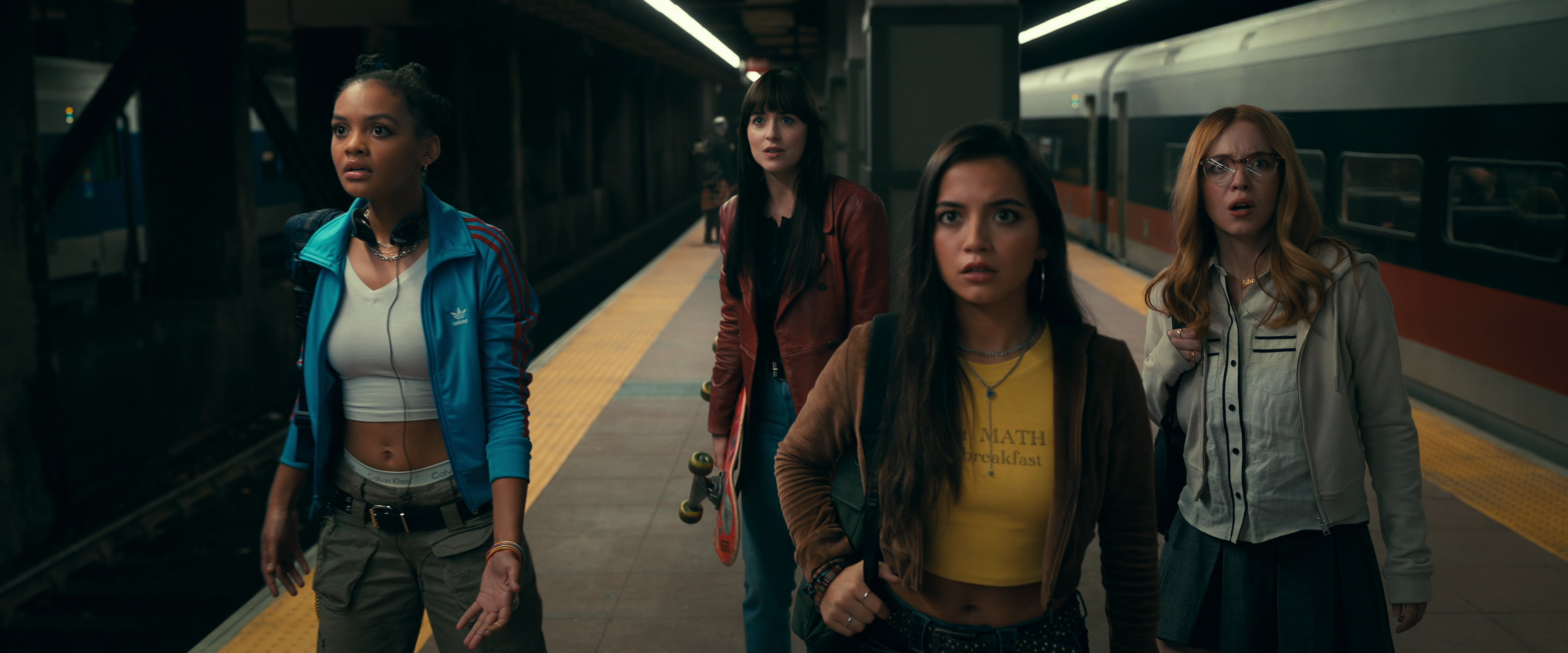 Four individuals stand on a subway platform, their faces not shown for privacy. One wears a blue jacket and grey pants, another a red top with a skateboard, the third a brown jacket and yellow top with text, and the fourth light-colored attire. An orange and white train is visible in the background, with ‘HEALTH & SAFETY’ text discernible, highlighting the urban commute environment.