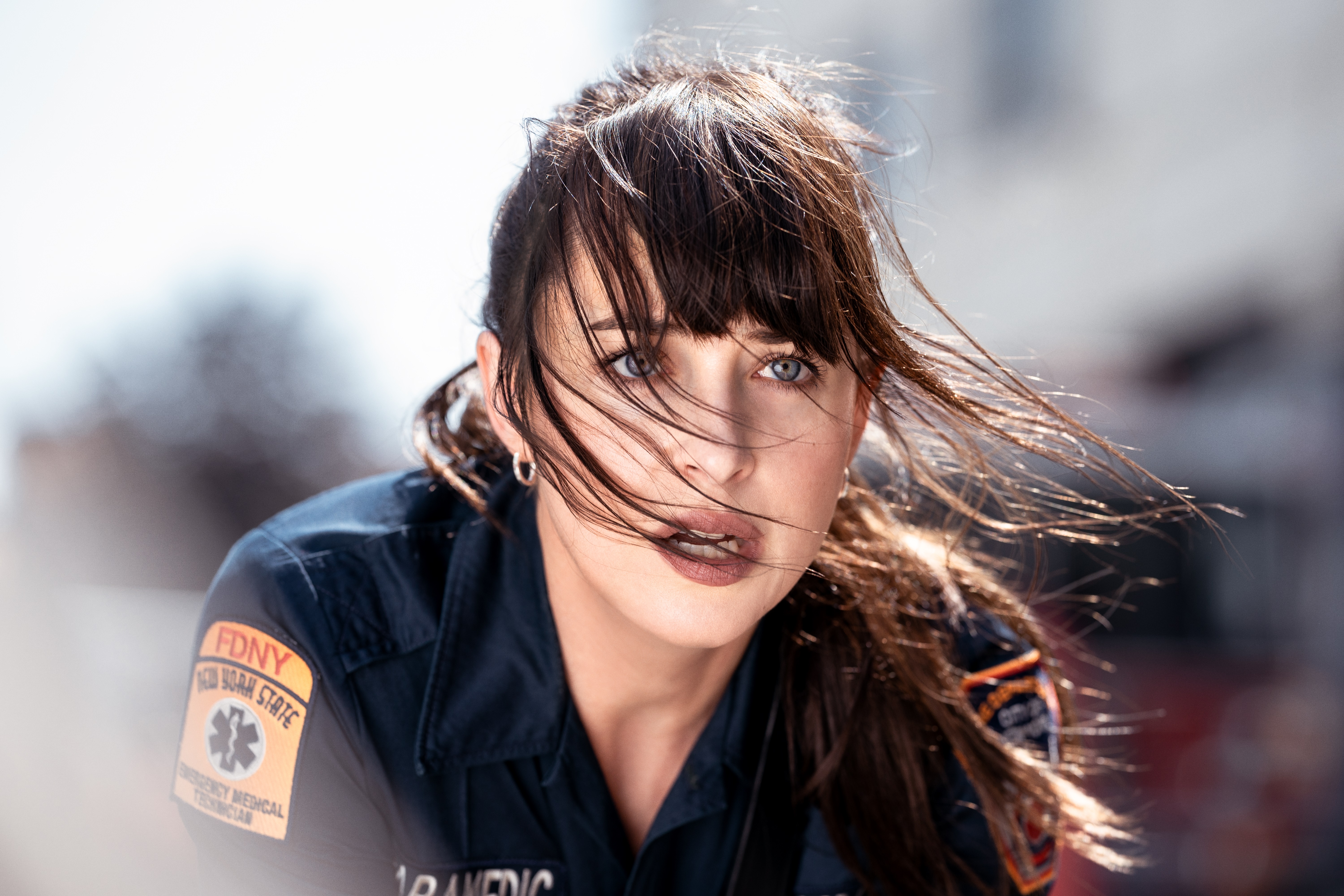 A person in a dark blue FDNY*EMS uniform stands with windblown hair, the blurred background suggesting an outdoor urban setting. Patches on the uniform, including the Star of Life symbol and an American flag, indicate their role in emergency medical services.