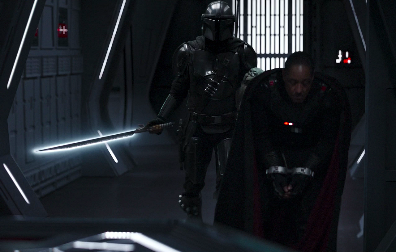 Djarin carries the ignited darksaber while escorting Moff Gideon onto the bridge.