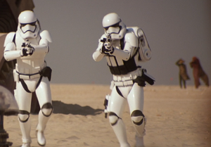 Two First Order stormtroopers with their weapons drawn move toward the viewer. Davan Marak and another individual are visible in the distance standing in the desert sands of Jakku.