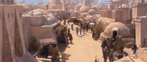 Rum Sleg, in bright red armor, and a human in green and black armor talk in the streets of Mos Espa on Tatooine.