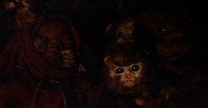A cluster of Ewoks stand in a darkened hut listening to stories. On the right, one holds Nippet and on the left stands Widdle Warrick.