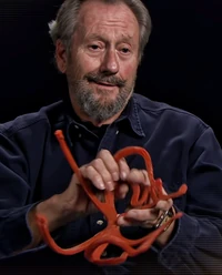 Richard Miller, a man with graying hair and a full beard and mustache, holds a red-orange rubber frame.