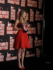 A young woman with blond, curly hair, in left profile, holds up an award. She is dressed in a red dress while standing on the red carpet.