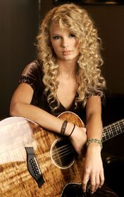 A young female with curly blond hair faces down at an acoustic guitar made of koa wood while a large microphone is placed close to her. She is wearing patterned, red dress.