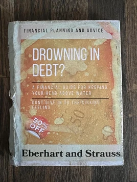 A standard hardback book titled “Drowning in debt? A financial guide to keeping your head above water” by Eberhart & Strauss. The cover is orange/tan with imagery of £50 bank notes, two markings show it was on offer. The entire book suffers extensive water damage resulting in the destruction of the spine and fading of the cover.
