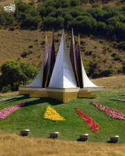 Six tall white and purple spires with gold trim rise from a three-pointed gold base, surrounded by radiating flower beds on a grassy mound.
