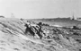 With bullets and artillery shells screaming overhead, Marines crawl along the beaches and dig into the soft volcanic ash for cover from the deadly fire. Note the geyser of water as a shell lands close to a landing craft headed into the beach.
