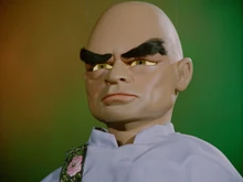 The Hood, wearing his light blue tunic, and with his eyes lit up yellow, frowns as he attempts to telepathically contact his brother Kyrano