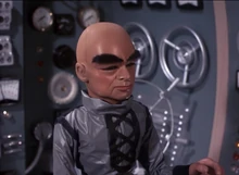 The Hood, wearing a grey wetsuit, surrounded by dials, flashing lights and other instruments