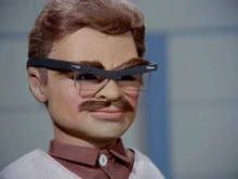 The Hood in disguise as Professor Langley; he is wearing a mask with realistic brown hair and a moustache, and a pair of glasses