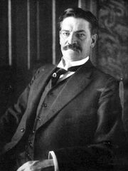 Photograph of a moustached middle-aged man in a dark suit and waistcoat, sitting in a chair while looking at the camera